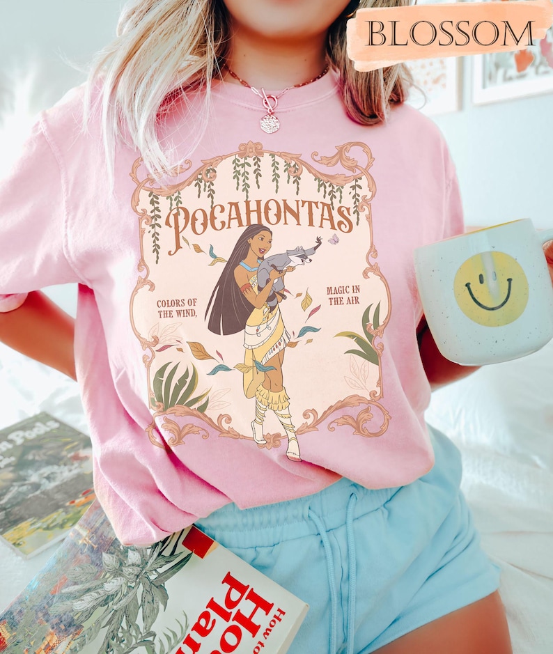 Op de afbeelding: Zachtroze t-shirt met een Pocahontas-afbeelding en de tekst "Pocahontas, Kleuren van de Wind, Magie in de Lucht". Het shirt wordt gedragen met een blauwe short en een witte mok met een gele smiley.