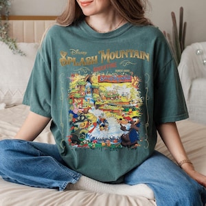 May include: A teal green t-shirt with a vintage-style graphic of Disney's Splash Mountain ride. The graphic features a cartoon illustration of the ride's characters and the text "Disney Splash Mountain Adventure Since 1989."