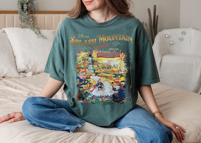 May include: A teal green t-shirt with a vintage-style graphic of Disney's Splash Mountain ride. The graphic features a cartoon illustration of the ride's characters and the text "Disney Splash Mountain Adventure Since 1989."