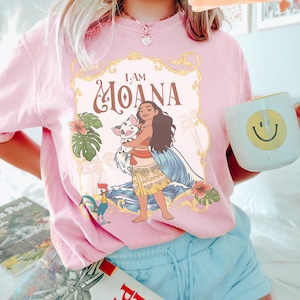 May include: Pink t-shirt featuring a graphic of Moana holding a pig, with the text "I AM MOANA". The design includes tropical flowers, a rooster, and ocean waves. The person is also holding a white mug with a yellow smiley face.