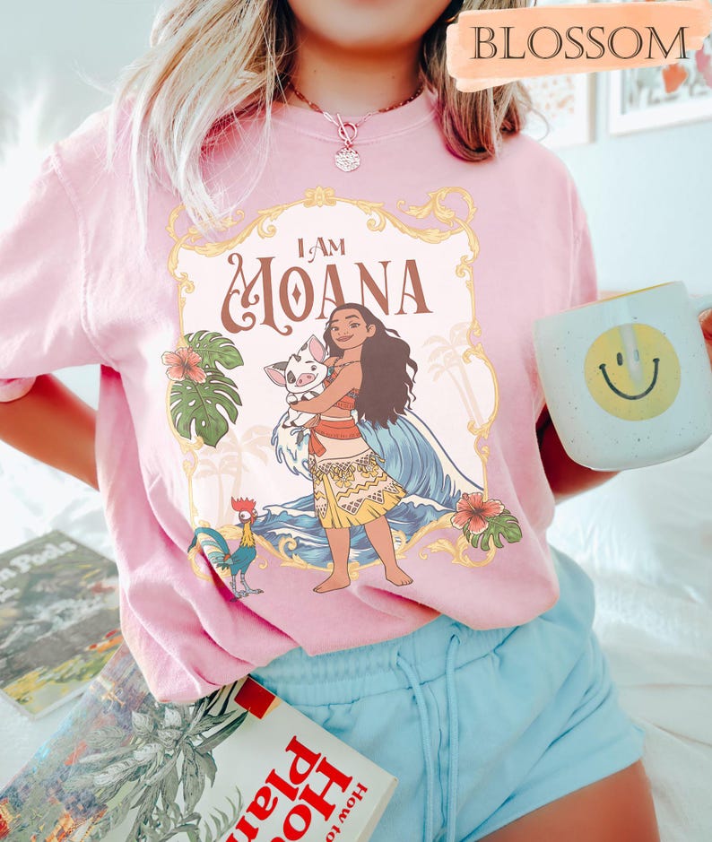 May include: Pink t-shirt featuring a graphic of Moana holding a pig, with the text "I AM MOANA". The design includes tropical flowers, a rooster, and ocean waves. The person is also holding a white mug with a yellow smiley face.