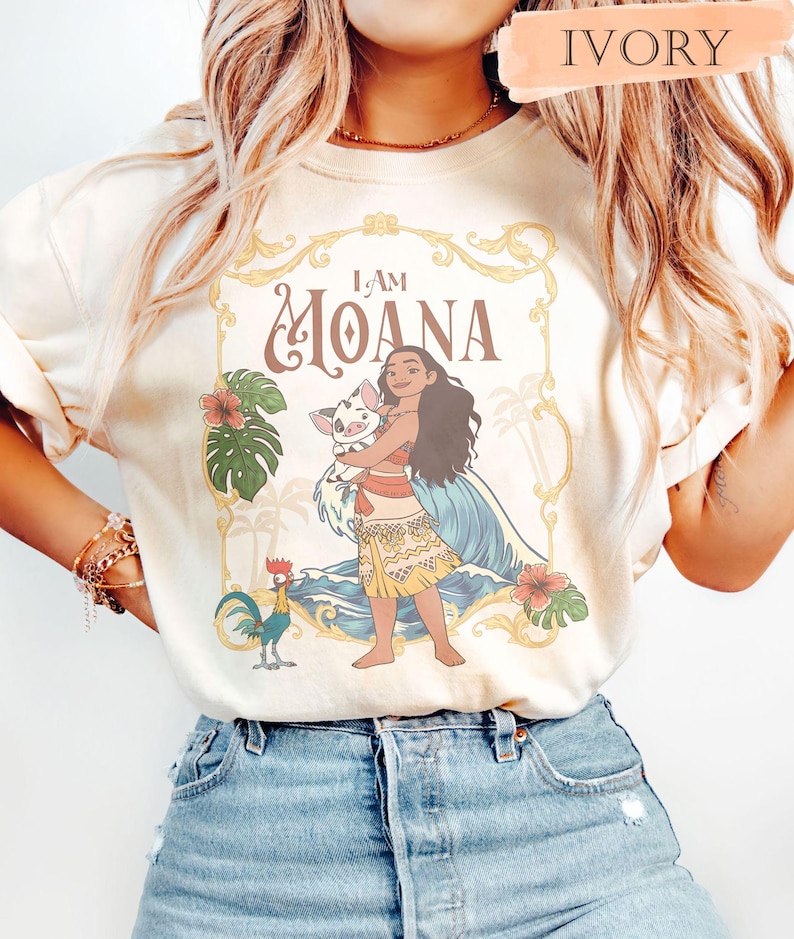 May include: Ivory-colored t-shirt featuring a graphic of Moana holding a pig, with the text "I AM MOANA." The design includes tropical flowers, palm trees, and a rooster. The shirt has short sleeves.