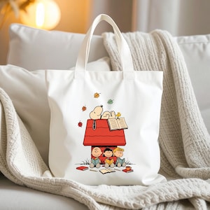 May include: White tote bag featuring a cartoon illustration of Snoopy and the Peanuts gang. Snoopy is on top of his red doghouse, reading a book, while the other characters are reading below. Autumn leaves are falling around them.