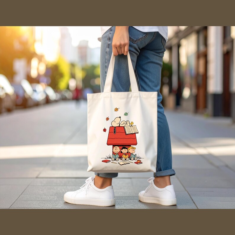 May include: A white tote bag featuring a cartoon illustration of Snoopy and friends reading in a red doghouse. The bag has white handles and is being held by a person wearing blue jeans and white sneakers.