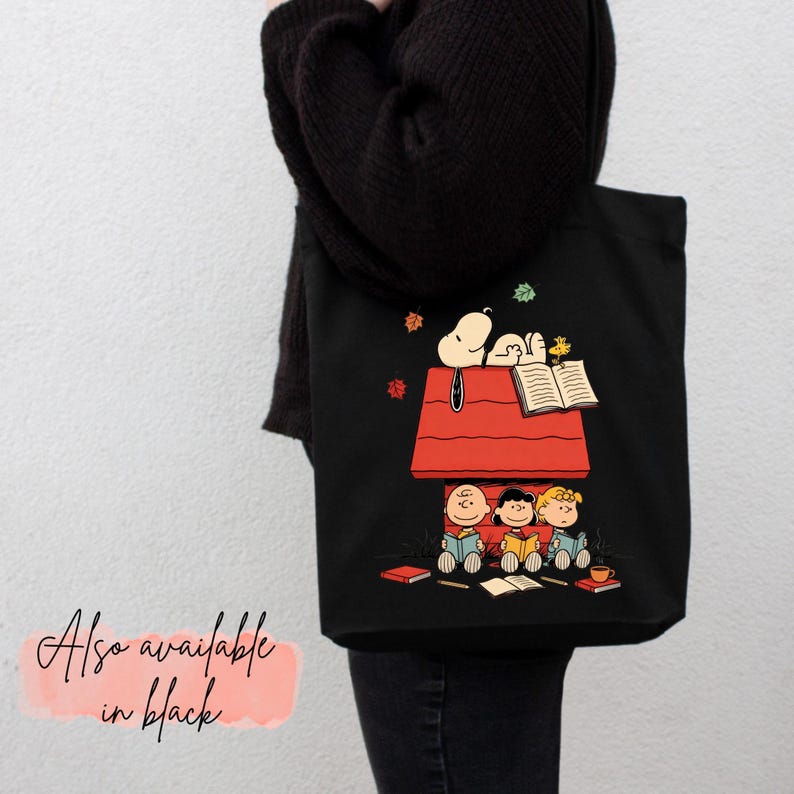 May include: Black tote bag featuring a Snoopy and the Peanuts gang design. Snoopy is on top of his red doghouse, reading a book, while Charlie Brown, Sally, and Lucy are reading below. The text "Also available in black" is in the bottom left corner.