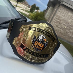 Pu&ograve; includere: Una cintura da campione nera con una grande placca centrale dorata raffigurante un'aquila, il testo 2025 CHAMPION, BMA LEAGUE e PAPI RICKY. La placca &egrave; decorata con piccole pietre trasparenti e intricati motivi in rilievo sui bordi.