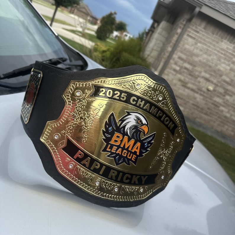 Pu&ograve; includere: Una cintura da campione nera con una grande placca centrale dorata raffigurante un'aquila, il testo 2025 CHAMPION, BMA LEAGUE e PAPI RICKY. La placca &egrave; decorata con piccole pietre trasparenti e intricati motivi in rilievo sui bordi.