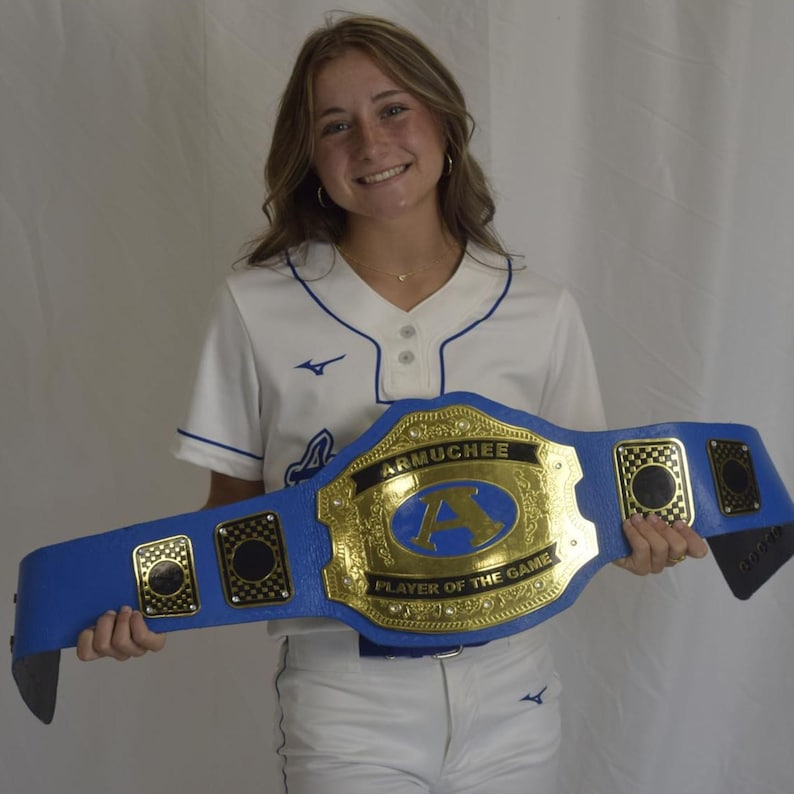 Pu&ograve; includere: Una cintura da campionato blu con una grande placca centrale dorata con il testo ARMUCHEE e PLAYER OF THE GAME, centrata con una lettera A blu. La cintura include placche laterali dorate pi&ugrave; piccole con motivi a scacchi neri.