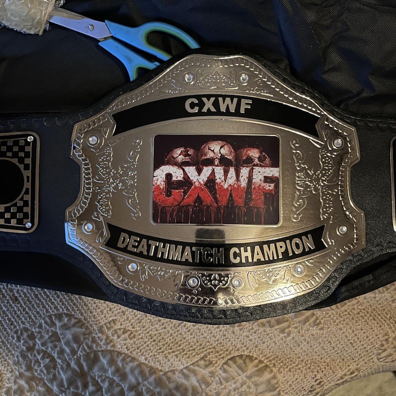 Pu&ograve; includere: Una cintura da campionato in pelle nera con una grande placca centrale in metallo argentato lucido. La placca mostra il testo CXWF e DEATHMATCH CHAMPION, insieme a un grafico centrale di tre teschi e il testo CXWF rosso sangue.