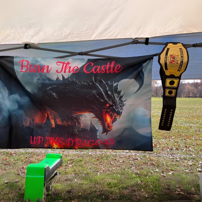 Pu&ograve; includere: Uno stendardo rettangolare raffigurante un drago nero con occhi rossi luminosi e bocca infuocata sopra un castello scuro. Il testo "Burn The Castle" e "UP THE DRAGONS" &egrave; stampato in rosso. Una cintura da campionato dorata pende a destra.