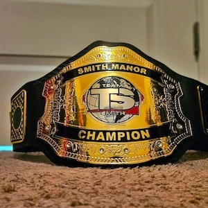 Pu&ograve; includere: Una cintura da campionato con una grande piastra centrale in metallo dorato lucido con accenti neri. La piastra mostra il testo SMITH MANOR in alto, CHAMPION in basso e un grafico centrale a forma di globo con le lettere TS e TEAM SMITH.