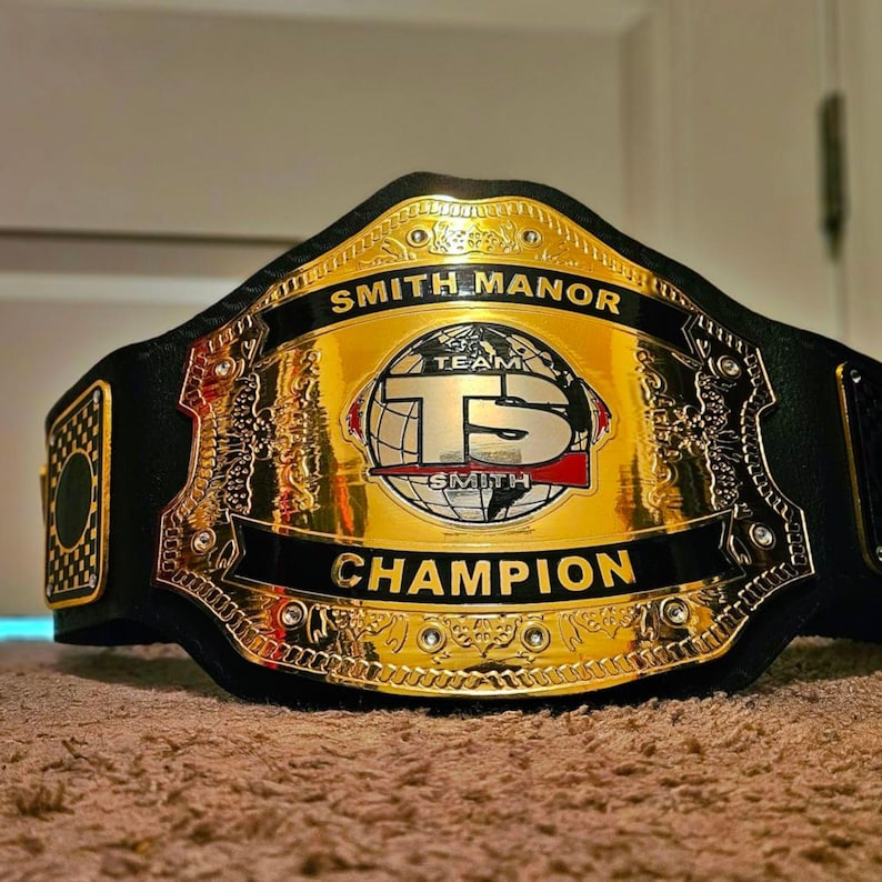 Pu&ograve; includere: Una cintura da campionato con una grande piastra centrale in metallo dorato lucido con accenti neri. La piastra mostra il testo SMITH MANOR in alto, CHAMPION in basso e un grafico centrale a forma di globo con le lettere TS e TEAM SMITH.