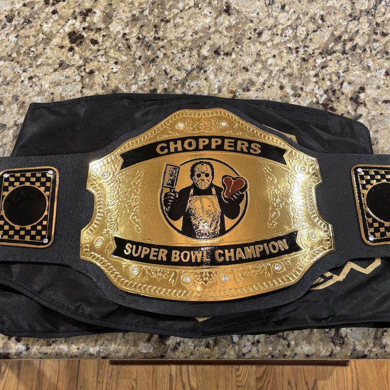 Pu&ograve; includere: Una cintura da campione nera con una grande placca centrale dorata che presenta l'illustrazione incisa di una figura con maschera da hockey che tiene in mano una mannaia e una bistecca. Il testo CHOPPERS e SUPER BOWL CHAMPION &egrave; su banner neri.