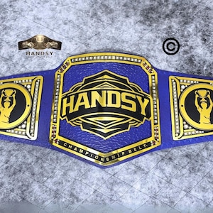 May include: A blue and gold championship belt with the word "HANDSY" in gold lettering on the front. The belt has two gold side plates with a silhouette of a person holding a trophy.