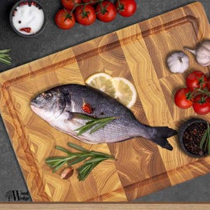 May include: A fresh fish with silver scales and dark fins is presented on a wooden chopping board. It's garnished with lemon, rosemary, tomatoes, garlic, and bowls of salt and peppercorns. The board features a geometric pattern.