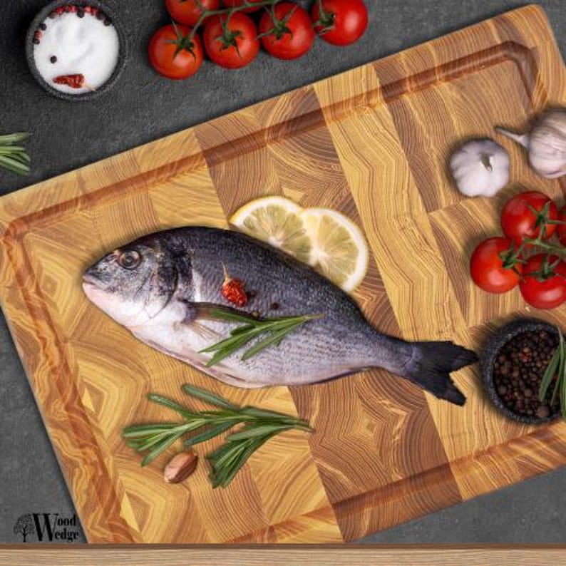 May include: A fresh fish with silver scales and dark fins is presented on a wooden chopping board. It's garnished with lemon, rosemary, tomatoes, garlic, and bowls of salt and peppercorns. The board features a geometric pattern.