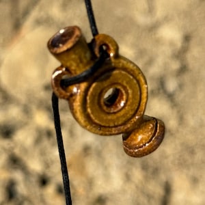 May include: A small, brown pendant with a spiral design and a cylindrical top, strung on a black cord. The pendant has a glossy finish and appears to be made of wood or a similar material. The design is intricate and detailed.