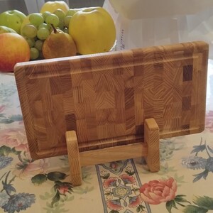 May include: A wooden chopping board with a butcher block pattern, supported by a wooden stand. The board is made of light-coloured wood and has a textured surface. A bouquet of blue hydrangeas and various fruits are in the background.