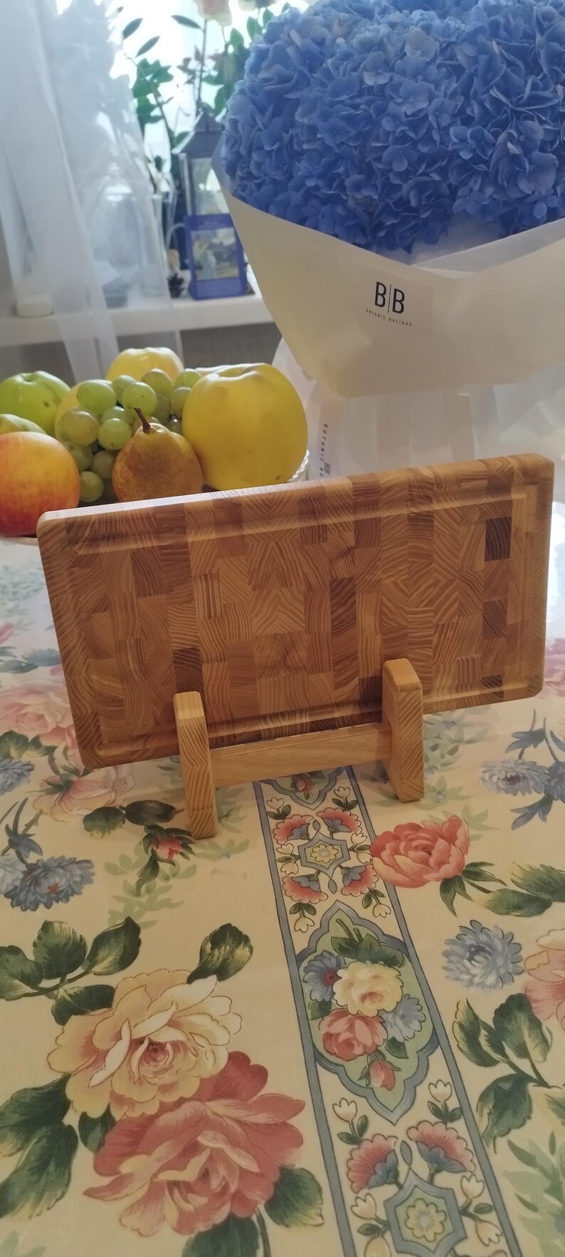 May include: A wooden chopping board with a butcher block pattern, supported by a wooden stand. The board is made of light-coloured wood and has a textured surface. A bouquet of blue hydrangeas and various fruits are in the background.