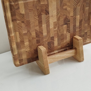 May include: A rectangular wooden chopping board with a butcher block design, supported by a wooden stand. The board is made of light brown wood and has a textured surface. The stand is also made of light brown wood.