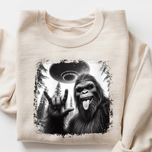 May include: A beige sweatshirt with a black and white graphic print of a Bigfoot giving the peace sign with a UFO in the background. The text "SAND" is in the upper right corner of the image.
