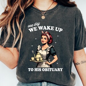 May include: A dark gray t-shirt with a vintage-style illustration of a woman holding a teapot and cups. The text on the shirt reads "one day WE WAKE UP TO HIS OBITUARY".