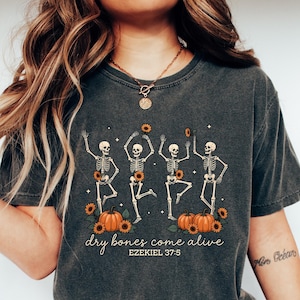 May include: Dark gray t-shirt featuring a dancing skeleton design with sunflowers and pumpkins. The text "dry bones come alive" and "EZEKIEL 37:5" is printed below the graphic. The shirt is paired with blue jeans and white sneakers.