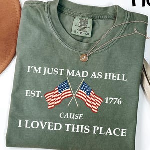 May include: Moss green t-shirt with the text "I'M JUST MAD AS HELL EST. 1776 CAUSE I LOVED THIS PLACE" and two crossed American flags. The shirt has a crew neck and short sleeves. A gold necklace is draped across the shirt.