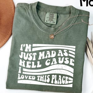 May include: A moss green t-shirt with the text "I'M JUST MAD AS HELL CAUSE I LOVED THIS PLACE" in white. The shirt has a crew neck and short sleeves. A gold necklace with a pendant is draped across the shirt.