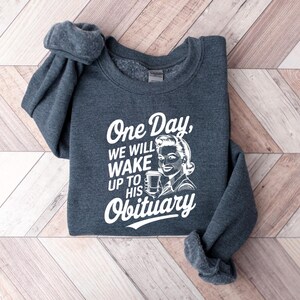 May include: A dark gray sweatshirt with a white graphic print that says "One Day, We Will Wake Up To His Obituary". The graphic features a retro-style illustration of a woman holding a cup of coffee.