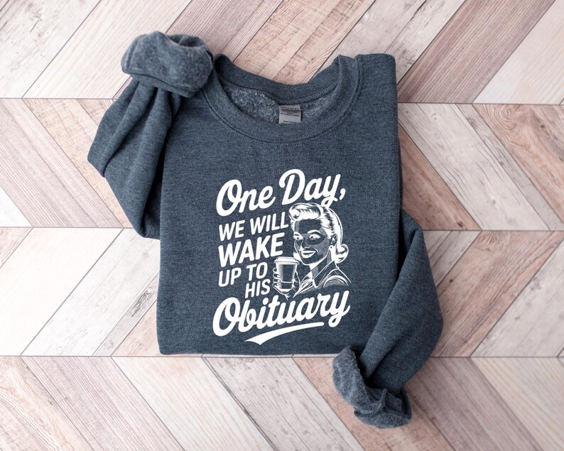 May include: A dark gray sweatshirt with a white graphic print that says "One Day, We Will Wake Up To His Obituary". The graphic features a retro-style illustration of a woman holding a cup of coffee.