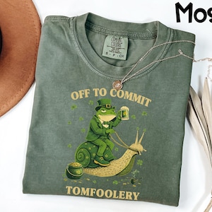 May include: Moss green t-shirt with the text "OFF TO COMMIT TOMFOOLERY". The shirt features a cartoon frog in a green suit riding a snail, holding a beer. A gold necklace is draped across the shirt.