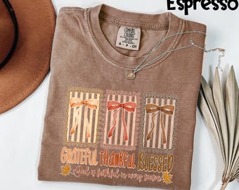 May include: Espresso-colored t-shirt with a graphic of three framed bows in orange, red, and brown. The text "GRATEFUL THANKFUL BLESSED" and "God is faithful in every season" are printed below the bows. A silver necklace is draped across the shirt.