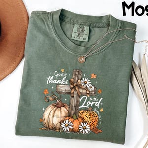 Puede incluir: Una camiseta verde musgo con un gr&aacute;fico de una cruz de madera, calabazas y margaritas. El texto "Give thanks to the Lord" est&aacute; impreso en la camiseta. Un collar plateado est&aacute; drapeado sobre la camiseta.