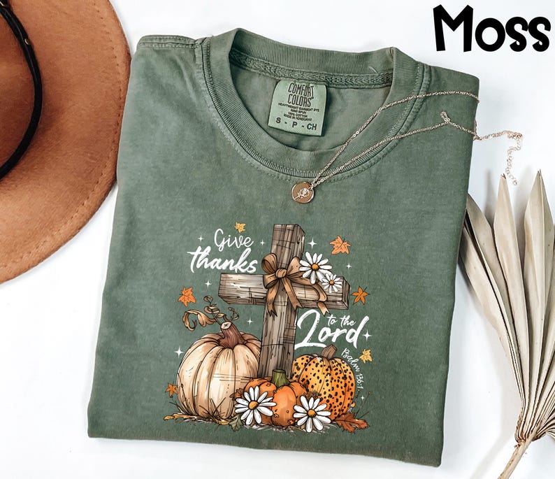 Puede incluir: Una camiseta verde musgo con un gr&aacute;fico de una cruz de madera, calabazas y margaritas. El texto "Give thanks to the Lord" est&aacute; impreso en la camiseta. Un collar plateado est&aacute; drapeado sobre la camiseta.