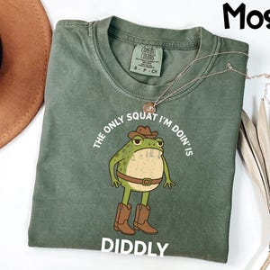 May include: A moss green t-shirt with a cartoon frog wearing a cowboy hat and boots. The text on the shirt reads "The only squat I'm doin' is DIDDLY". A gold necklace is draped across the shirt.