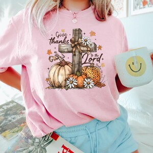 Puede incluir: Camiseta rosa de manga larga con un dise&ntilde;o gr&aacute;fico que presenta una cruz de madera, calabazas, margaritas y el texto "Give thanks to the Lord". El dise&ntilde;o incluye hojas de oto&ntilde;o y el vers&iacute;culo b&iacute;blico Salmo 86:12. Se sostiene una taza blanca con una carita sonriente.
