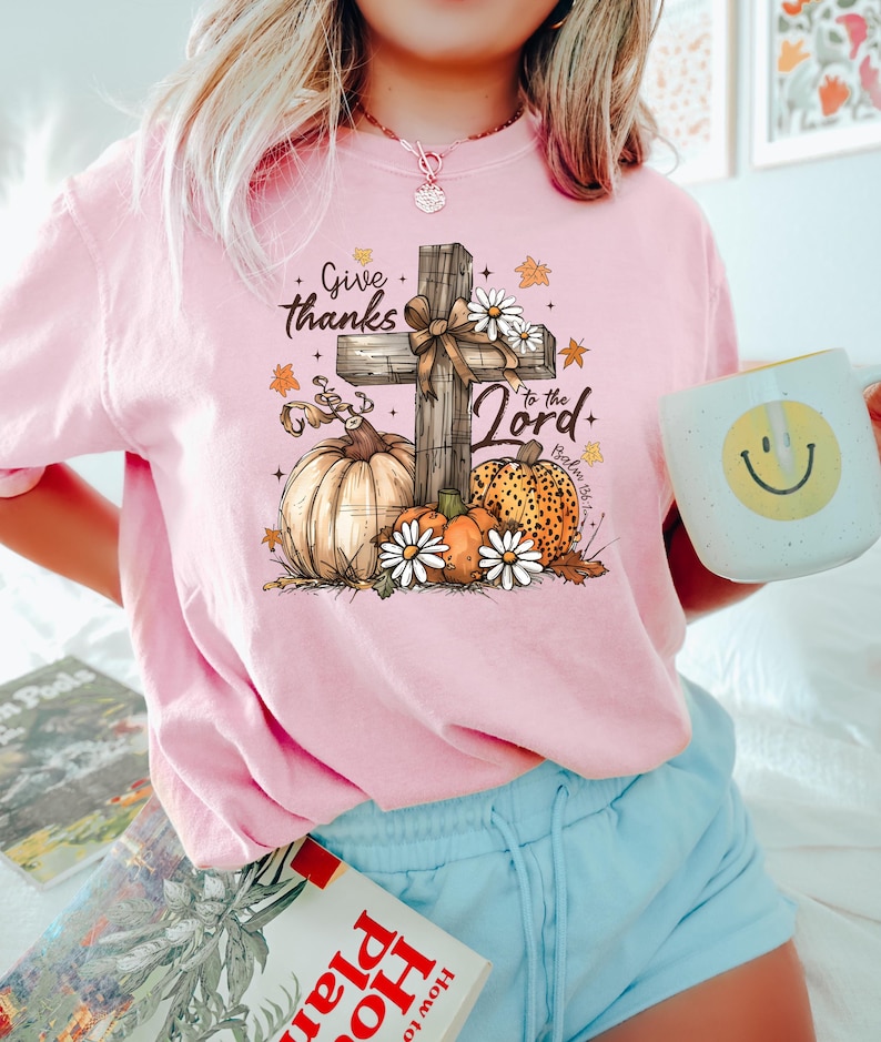 Puede incluir: Camiseta rosa de manga larga con un dise&ntilde;o gr&aacute;fico que presenta una cruz de madera, calabazas, margaritas y el texto "Give thanks to the Lord". El dise&ntilde;o incluye hojas de oto&ntilde;o y el vers&iacute;culo b&iacute;blico Salmo 86:12. Se sostiene una taza blanca con una carita sonriente.