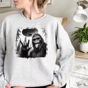 May include: Gray sweatshirt with a black and white graphic of a Bigfoot giving the peace sign with a UFO in the background.