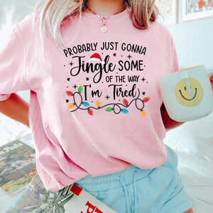 Puede incluir: Camiseta rosa con el texto "Probably Just Gonna Jingle Some of the Way - I'm Tired" en escritura negra y una guirnalda de luces navide&ntilde;as. Se sostiene una taza blanca con una carita sonriente amarilla.