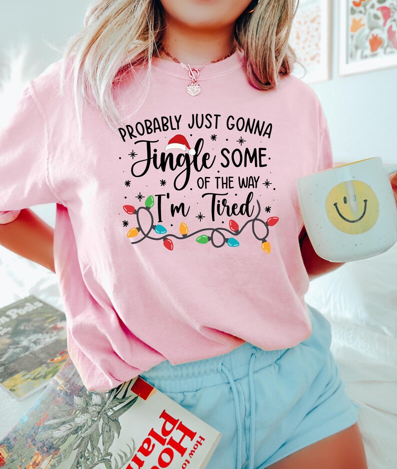 Puede incluir: Camiseta rosa con el texto "Probably Just Gonna Jingle Some of the Way - I'm Tired" en escritura negra y una guirnalda de luces navide&ntilde;as. Se sostiene una taza blanca con una carita sonriente amarilla.