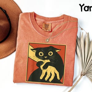 May include: A coral-colored t-shirt featuring a graphic of a black cat biting a yellow hand, set against a red background. The shirt has a crew neck and a small tag. A gold necklace and a brown hat are also visible.