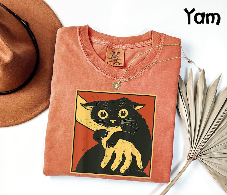 May include: A coral-colored t-shirt featuring a graphic of a black cat biting a yellow hand, set against a red background. The shirt has a crew neck and a small tag. A gold necklace and a brown hat are also visible.