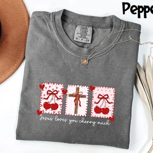May include: A gray t-shirt with a graphic design featuring three rectangular patches. The patches display a cross, cherries, and the text "Jesus loves you cherry much". The shirt has a crew neck and short sleeves.