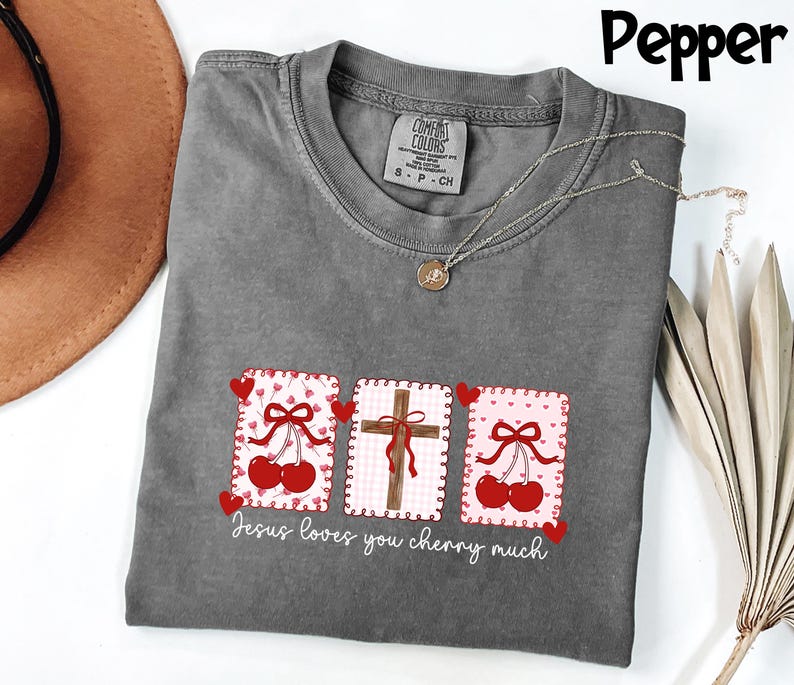 May include: A gray t-shirt with a graphic design featuring three rectangular patches. The patches display a cross, cherries, and the text "Jesus loves you cherry much". The shirt has a crew neck and short sleeves.