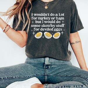May include: Dark gray t-shirt with the text "I wouldn't do a Lot for turkey or ham + but I would do + some sketchy stuff + for deviled eggs +" and a graphic of three deviled eggs. The shirt is paired with blue jeans and white shoes.