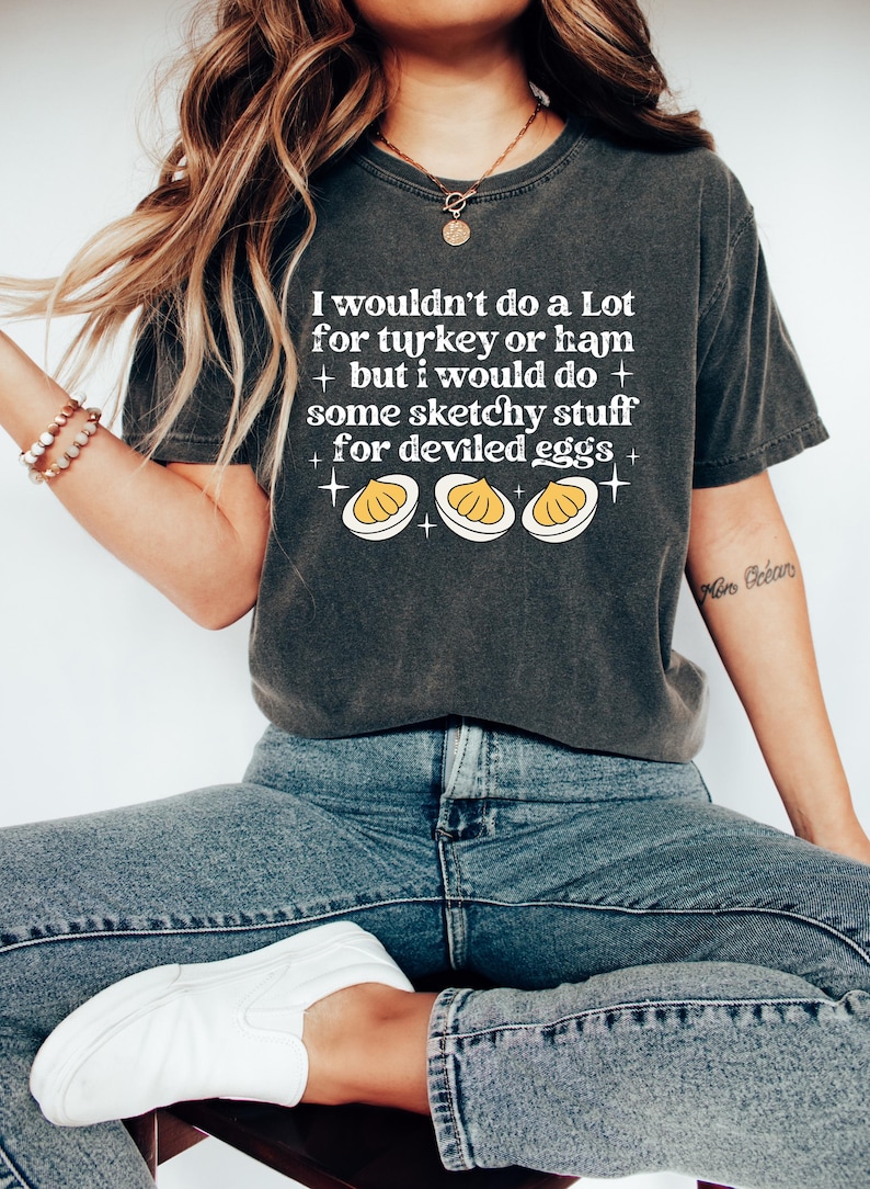 May include: Dark gray t-shirt with the text "I wouldn't do a Lot for turkey or ham + but I would do + some sketchy stuff + for deviled eggs +" and a graphic of three deviled eggs. The shirt is paired with blue jeans and white shoes.