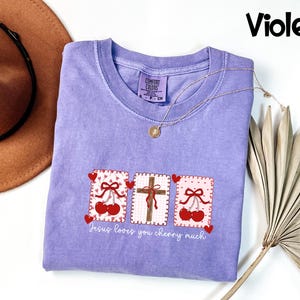 May include: A violet-colored t-shirt with a crew neck and short sleeves. The shirt features three rectangular designs with cherries, hearts, and a cross. The text "Jesus loves you cherry much" is printed below the designs. A gold necklace is draped on the shirt.