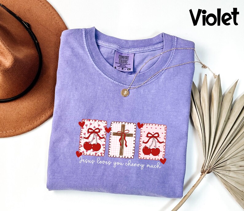 May include: A violet-colored t-shirt with a crew neck and short sleeves. The shirt features three rectangular designs with cherries, hearts, and a cross. The text "Jesus loves you cherry much" is printed below the designs. A gold necklace is draped on the shirt.