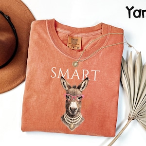 May include: A coral-colored t-shirt with the word "SMART" printed in white. The shirt features a graphic of a donkey wearing pink glasses and a pearl necklace. A gold necklace and a brown hat are also visible.